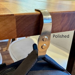 K&ouml;nnte beinhalten: Ein polierter silberner Haken zum Aufh&auml;ngen von Taschen oder anderen Gegenst&auml;nden unter einem Tisch. Der Haken ist mit zwei Schrauben an der Unterseite des Tisches befestigt. Der Text "Polished" ist auf dem Tisch sichtbar.