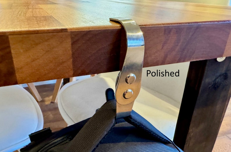 K&ouml;nnte beinhalten: Ein polierter silberner Haken zum Aufh&auml;ngen von Taschen oder anderen Gegenst&auml;nden unter einem Tisch. Der Haken ist mit zwei Schrauben an der Unterseite des Tisches befestigt. Der Text "Polished" ist auf dem Tisch sichtbar.