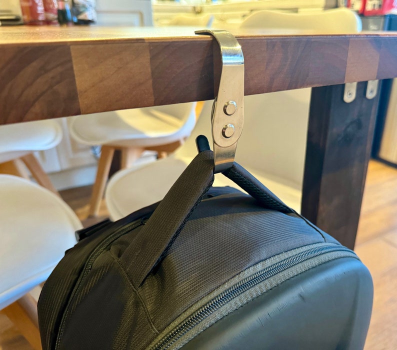 K&ouml;nnte beinhalten: Ein silberner Haken aus Metall h&auml;ngt an einem Holztisch und h&auml;lt einen schwarzen Rucksack.