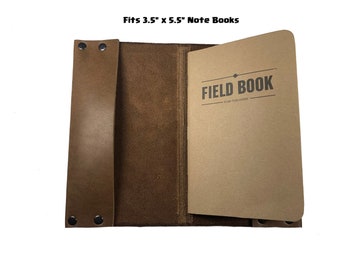 May include: Brown leather notebook cover with a flap and a strap. The cover fits a 3.5" x 5.5" notebook. The notebook is a Field Book brand notebook with the text "FIELD BOOK" printed on the cover.