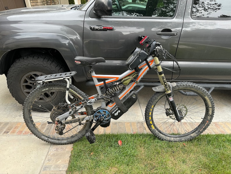 May include: A silver and orange electric mountain bike with a black battery pack and a black seat. The bike has a front suspension fork and a rear suspension. The bike is parked next to a gray pickup truck.