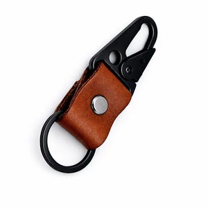 May include: A brown leather keyring featuring a matte black metal carabiner clip and a matching black circular ring. The leather strap is secured with a silver-toned metal snap button, connecting the carabiner to the circular ring.