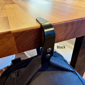 K&ouml;nnte beinhalten: Schwarzer Metallhaken zum Aufh&auml;ngen einer Tasche oder anderer Gegenst&auml;nde an einem Tisch oder Schreibtisch. Der Haken ist an der Unterseite des Tisches befestigt und hat zwei schwarze Kn&ouml;pfe. Der Text "Black" ist auf dem Tisch geschrieben.
