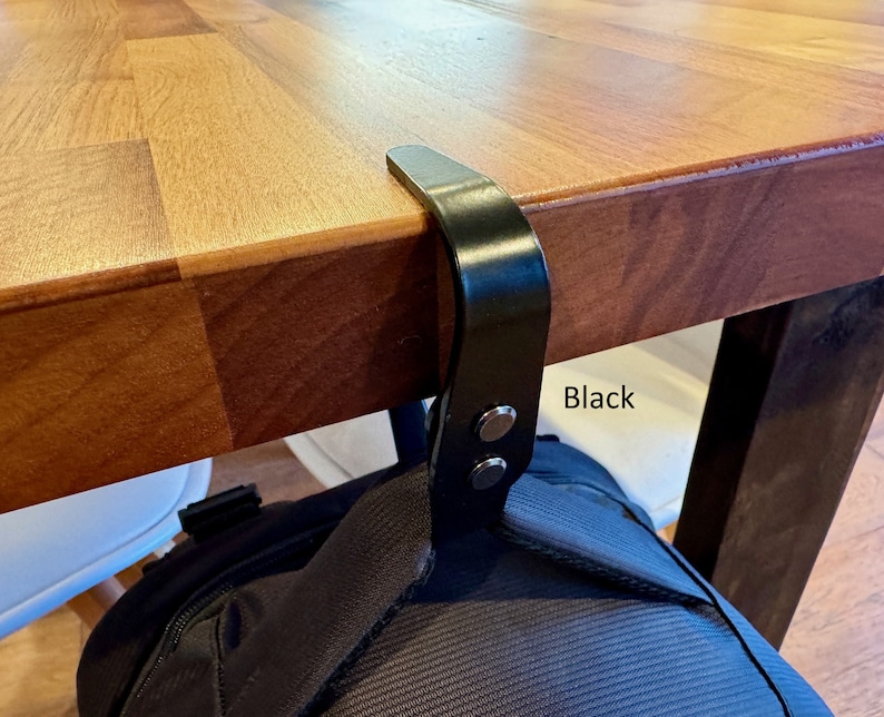 K&ouml;nnte beinhalten: Schwarzer Metallhaken zum Aufh&auml;ngen einer Tasche oder anderer Gegenst&auml;nde an einem Tisch oder Schreibtisch. Der Haken ist an der Unterseite des Tisches befestigt und hat zwei schwarze Kn&ouml;pfe. Der Text "Black" ist auf dem Tisch geschrieben.