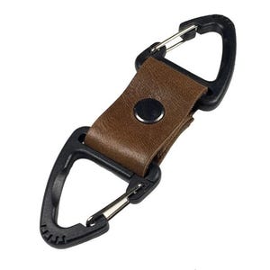May include: A black carabiner with a brown leather strap and a silver clasp. The strap is secured with a black snap. This item is likely used for attaching items.