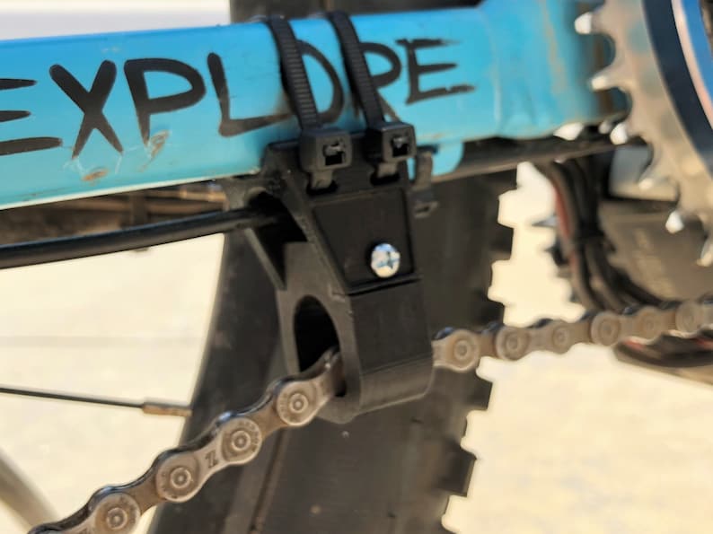 Peut inclure: Gros plan d'un guide-cha&icirc;ne de v&eacute;lo noir fix&eacute; &agrave; un cadre de v&eacute;lo bleu avec le mot "EXPLORE" &eacute;crit en peinture noire. Le guide-cha&icirc;ne a une vis et un petit trou pour fixer un serre-c&acirc;ble.