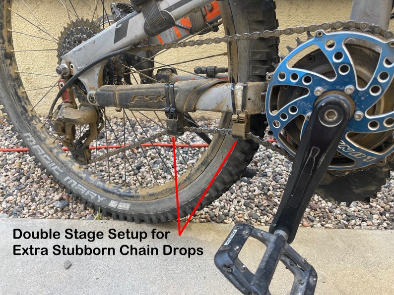 Peut inclure: Gros plan sur une cha&icirc;ne de v&eacute;lo avec un syst&egrave;me &agrave; double &eacute;tage pour les chutes de cha&icirc;ne les plus tenaces. La cha&icirc;ne est sur un d&eacute;railleur arri&egrave;re avec un plateau bleu et argent. La cha&icirc;ne est reli&eacute;e &agrave; un bras de manivelle noir avec une p&eacute;dale noire.