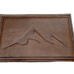 May include: A brown leather patch featuring an embossed mountain range design. The rectangular patch has a raised border and a textured surface. The mountain range is in the center.