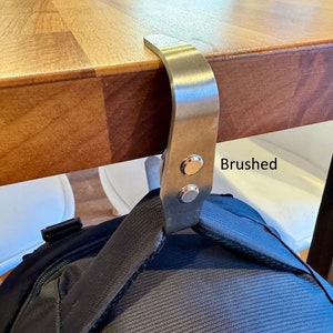K&ouml;nnte beinhalten: Ein geb&uuml;rsteter Edelstahlhaken, der unter einem Holztisch montiert ist und einen schwarzen Rucksack h&auml;lt.