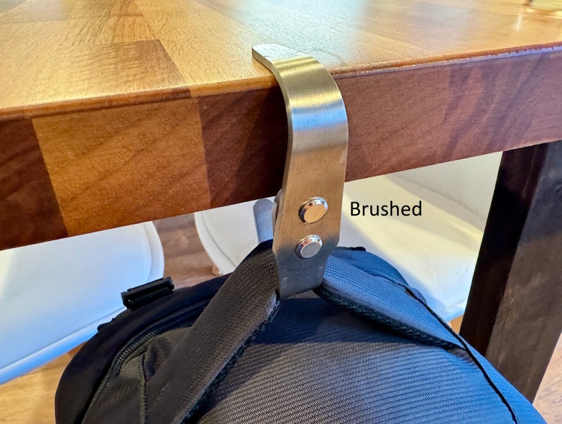 K&ouml;nnte beinhalten: Ein geb&uuml;rsteter Edelstahlhaken, der unter einem Holztisch montiert ist und einen schwarzen Rucksack h&auml;lt.