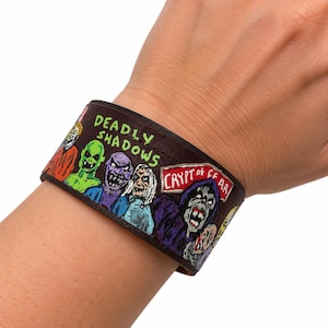 May include: A dark brown leather cuff bracelet featuring hand-painted illustrations of various monsters and ghouls. The text "DEADLY SHADOWS" is printed in green, and a red sign displays the text "CRYPT OF FEAR" alongside colorful creature designs.