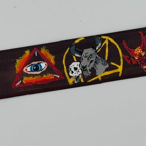 May include: A dark brown leather belt featuring hand-painted designs including a flaming red triangle with a blue eye, a grey goat head with a skull inside a yellow pentagram, and a red horned figure with a yellow star on its forehead.