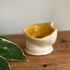May include: A small, handmade ceramic bowl with a unique, twisted design. The interior is bright yellow, contrasting with the off-white exterior. The bowl sits on a wooden surface, showcasing its handcrafted nature.