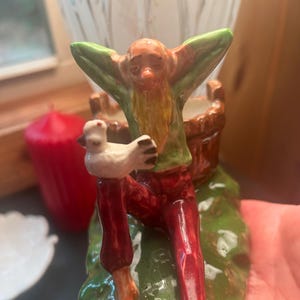 May include: A ceramic figurine of a seated gnome with a long beard, wearing a green shirt and red pants. The gnome holds a white dove. The figurine is perched on a green base with a brown bucket.
