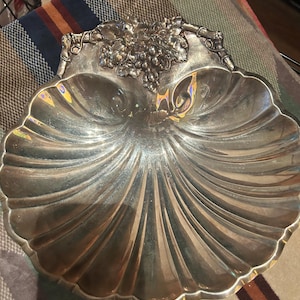 May include: Silver-toned, shell-shaped serving dish with a scalloped edge. The dish features a detailed grape cluster design at the top. The interior has a reflective, polished surface. A decorative piece for serving or display.