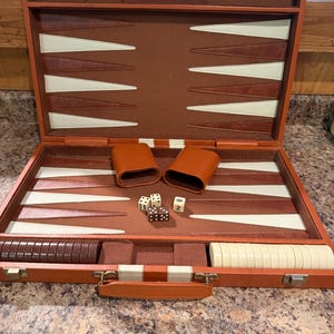 May include: A brown backgammon set in a briefcase-style case with a brown felt interior featuring cream and brown triangular points. The set includes two brown leather dice cups, two cream dice, one brown die, and a set of brown and cream checkers.