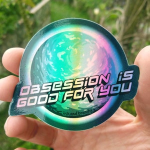 May include: A holographic sticker with a swirling galaxy design and the text "Obsession is good for you" in white letters. The sticker is on a dark green background and has the text "ink and brandy" in white letters at the bottom.