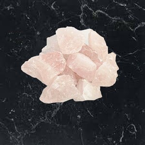 May include: A cluster of raw, pale pink rose quartz crystals. The crystals are of varying sizes and shapes, with a slightly translucent quality. The background is a dark, marbled surface.