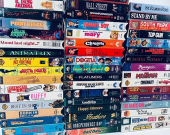 May include: A large collection of assorted VHS movie tapes stacked in three vertical columns. The spines display various film titles including Willow, Wall Street, Terminator 2, South Park, Clueless, Ace Ventura, and Back to the Future in colorful fonts.