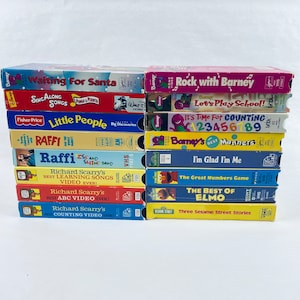 May include: A stack of 12 VHS tapes featuring popular children's shows, including Barney, Sesame Street, Raffi, and Richard Scarry. The tapes are arranged vertically, with the titles facing forward. The tapes are all in good condition and appear to be from the 1990s.