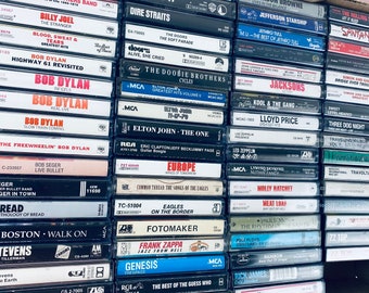 May include: A collection of vintage cassette tapes featuring various artists and albums, including Bob Dylan, The Eagles, Elton John, and Led Zeppelin. The tapes are arranged in a vertical stack, with the titles and artist names visible on the front of each tape.