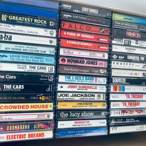 May include: A close-up view of a collection of cassette tapes, stacked vertically. The tapes are a mix of different artists and genres, including 80's rock, pop, and new wave. Some of the artists featured include Eurythmics, Falco, The Cars, and The J. Geils Band.