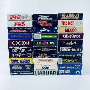May include: A stack of VHS tapes featuring movie titles like "American Pie," "The Fast and the Furious," and "Moulin Rouge." The tapes are in their original packaging, with colorful labels and movie titles prominently displayed. The collection includes a variety of genres.