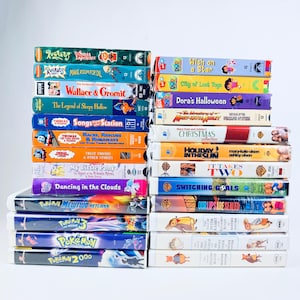May include: A collection of twenty-five VHS tapes stacked in two columns. The spines feature various titles including Rugrats, Dora the Explorer, Thomas the Tank Engine, My Little Pony, Pokemon, Mary-Kate and Ashley, and The Tale of Peter Rabbit.
