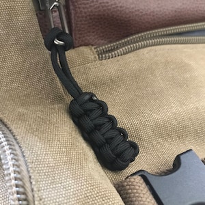 Puede incluir: Primer plano de un tirador de cremallera de paracord negro unido a una bolsa de lona marrón. El tirador de cremallera tiene un nudo en el extremo y está unido a una hebilla de plástico negra.