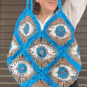May include: A blue, white, and brown crocheted tote bag with a geometric eye-like pattern. The bag has two shoulder straps and is made of a woven material. The bag is held up by the straps.