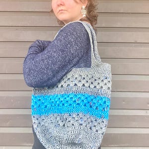 May include: A crocheted tote bag with a blue and gray color scheme. The bag has a wide, flat base and two long straps.