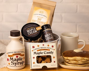 May include: A white metal bucket filled with maple syrup, maple cream, maple sugar, and maple candy. The bucket is surrounded by a brown paper bag with the text "Gluten-Free Oat Buttermilk" and a stack of pancakes with butter and syrup.