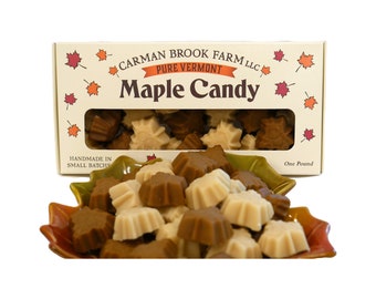 May include: A box of maple candy in the shape of maple leaves. The box is white with brown lettering that reads "Carman Brook Farm LLC Pure Vermont Maple Candy". The box is sitting on a table with a bowl of maple candy in front of it. The candy is a mix of light and dark brown.