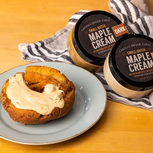 May include: Two jars of maple cream, one labeled "Dark" and the other "Light", both with the text "Carman Brook Farm Small Batch Maple Cream" and "No Preservatives, No Additives". A donut with maple cream frosting is on a blue plate.
