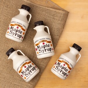 May include: Four white plastic bottles of 100% pure Vermont maple syrup with a brown and orange label. The label reads "Carman Brook Farm LLC" and "100% Pure Vermont Product". The bottles are on a brown burlap surface.