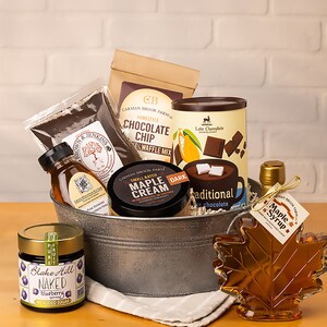 May include: A metal bucket filled with a variety of gourmet food items, including maple syrup, chocolate chips, waffle mix, maple cream, honey, and blueberry jam. The bucket is sitting on a wooden table with a white brick wall in the background.