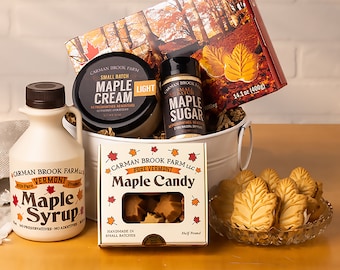 May include: A white metal bucket filled with maple syrup, maple cream, maple sugar, maple candy, and maple leaf cookies. The bucket is surrounded by fall-themed packaging and a white brick background.