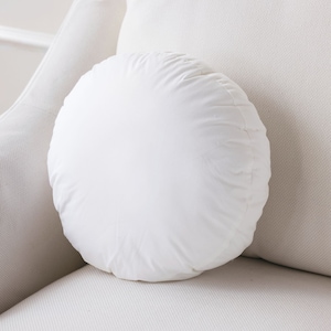 May include: A white round pillow with a gathered seam around the edge. The pillow is sitting on a white couch.
