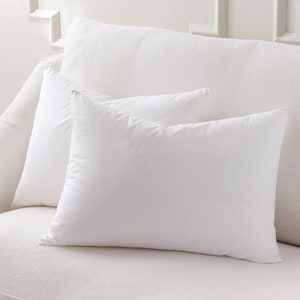 May include: Two white rectangular pillows rest on a white sofa. The pillows are fluffy and appear to be made of a soft material. The background is a white wall with decorative molding, creating a clean and minimalist aesthetic.