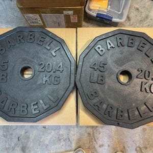 May include: Two black 45 lb (20.4 kg) weight plates with an octagonal shape and the word "BARBELL" printed around the edge.