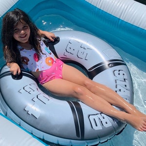 Könnte beinhalten: Ein junges Mädchen in einem rosa-weißen Badeanzug sitzt in einem grau-schwarzen aufblasbaren Ring in einem blauen Pool. Der Ring hat die Aufschrift "Get Wet" und "Splash" aufgedruckt.