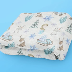 Op de afbeelding: Een wit fleece deken met een aquarelprint van verschillende Frozen-karakters, waaronder Elsa, Olaf en rendieren. De print bevat ook sneeuwvlokken en dennenbomen.