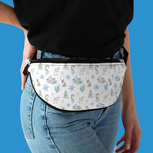 Op de afbeelding: Een witte heuptas met een zwarte rits en zwarte bies. De heuptas heeft een herhalend patroon van winterse illustraties, waaronder sneeuwvlokken, dennenbomen, rendieren en een persoon op een slee.