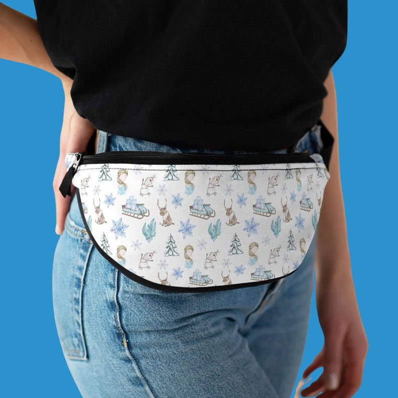Op de afbeelding: Een witte heuptas met een zwarte rits en zwarte bies. De heuptas heeft een herhalend patroon van winterse illustraties, waaronder sneeuwvlokken, dennenbomen, rendieren en een persoon op een slee.