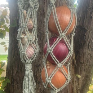 May include: Two sage green macrame produce hammocks hanging from metal hooks. The left hammock holds three heads of garlic, while the right hammock contains three onions, including one red onion and two yellow onions, finished with tassels.