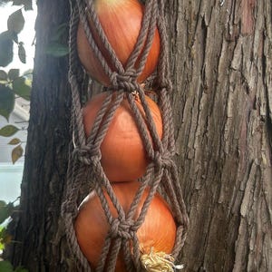 May include: A brown macrame rope hanger featuring a knotted net design that holds three round yellow onions in a vertical stack, finished with a looped top for hanging and a decorative tassel at the bottom end of the structure.