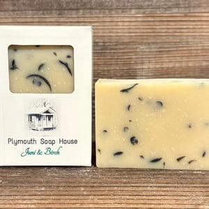 May include: A bar of soap and its white box. The soap is light yellow with dark specks. The box has a window showing the soap and a label with a house illustration and the text "Plymouth Soap House Juni & Birch."