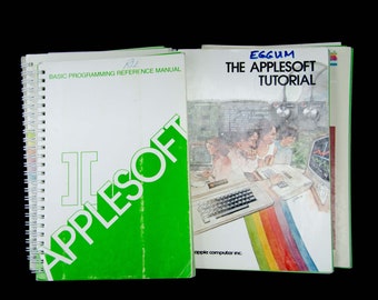 May include: A stack of vintage Apple II computer manuals, including the AppleSoft BASIC Programming Reference Manual and the AppleSoft Tutorial. The manuals are bound in white and green with the Apple logo on the cover.