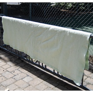 May include: A light green towel draped over a metal railing. The towel is made of a soft, absorbent material and is hanging outside. The background includes a chain-link fence and a brick surface.