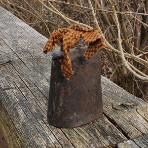 Puede incluir: Una campana de metal desgastado con base acampanada, adornada con tela a cuadros naranja y negro. La campana descansa sobre una superficie de madera desgastada. El fondo presenta ramas naturales. La campana es probablemente de hierro.